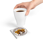Handheld white cup above square ceramic coaster printed with Byte & Bark Brew® circular dog logo