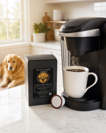 Steam rising from white mug beside single-serve brewer, Byte & Bark Brew® pod box, dog in background