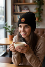 Person wearing a black knit beanie with embroidered dog patch, holding a steaming coffee cup